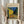 May include: Person in beige clothing holding framed Van Gogh reproduction of Cafe Terrace at Night, showing vibrant yellow cafe building against starry blue sky with outdoor dining tables on cobblestone street. Dried pampas grass visible to left, neutral curtained background. Light wood frame with cream matting displays the iconic 1888 painting with rich blues, yellows, and warm lighting depicting evening street scene.
