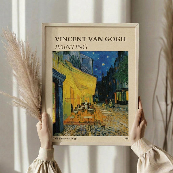 May include: Person in beige clothing holding framed Van Gogh reproduction of Cafe Terrace at Night, showing vibrant yellow cafe building against starry blue sky with outdoor dining tables on cobblestone street. Dried pampas grass visible to left, neutral curtained background. Light wood frame with cream matting displays the iconic 1888 painting with rich blues, yellows, and warm lighting depicting evening street scene.