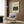 May include: Henri Rousseau jungle painting poster displayed on wood paneled wall above beige upholstered armchair with geometric patterned pillow. Modern reading corner with brass floor lamp, dark side table with open book, and white curtains. Poster shows lush green tropical foliage with hidden lion hunting scene in cream colored frame.