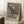 May include: Framed poster print featuring Edvard Munch's The Scream artwork in black and white, showing the iconic figure with hands to face on a bridge with two silhouettes in background. Bold text below reads "THE SCREAM - A MASTERPIECE OF EXPRESSIONISM" with artist name and date. Light wooden frame leaning against white wall corner next to cozy armchair and wooden side table with coffee cup on white textured rug.