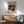 May include: Japanese woodblock print artwork displayed in modern living room with wooden sideboard, mint green upholstered chair, and white table lamp. The framed poster shows a figure in colorful robes against dark snowy background with Japanese text above. Warm sunlight streams across white walls creating geometric shadows. Mid-century modern furniture in natural wood tones complements the traditional art style.