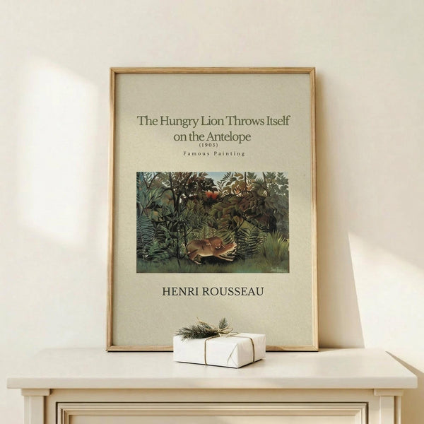 May include: Framed art print in light wood frame showing Henri Rousseau's jungle painting with lion attacking antelope amid lush green foliage. Print has cream background with black text identifying the 1905 artwork. Displayed on light wooden surface with small white wrapped gift box beside it against neutral wall.