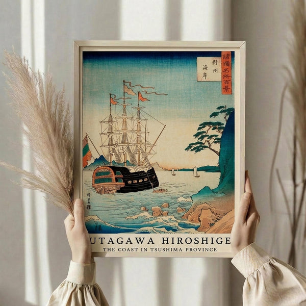 May include: Person in cream sweater holding a framed reproduction of a vintage Japanese woodblock print showing a large sailing ship with multiple masts near a rocky coastline with waves. The artwork features blue and teal waters, beige cliffs, and traditional Japanese text in red cartouches. Dried pampas grass visible on the left side, white curtains in background.