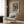 May include: Framed poster of Edvard Munch's The Scream artwork displayed on wood-paneled wall above modern cream armchair with geometric pillow. Black and white expressionist print shows figure with hands on face on bridge. Room features brass floor lamp, dark round side table with open book, and neutral tones throughout.