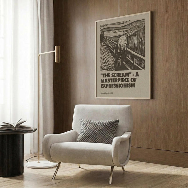 May include: Framed poster of Edvard Munch's The Scream artwork displayed on wood-paneled wall above modern cream armchair with geometric pillow. Black and white expressionist print shows figure with hands on face on bridge. Room features brass floor lamp, dark round side table with open book, and neutral tones throughout.