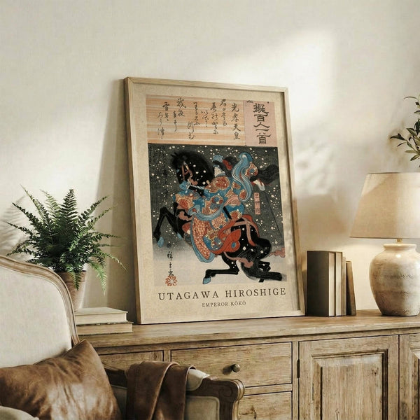 May include: Framed Japanese ukiyo-e woodblock print displayed on rustic wooden sideboard in neutral-toned living room. Print shows two figures in colorful robes amid falling snow on dark background, with Japanese text above. Scene includes fern plant, ceramic vase, books, and beige upholstered furniture.