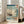 May include: Framed Japanese woodblock print reproduction leaning against white wall next to beige sofa in bright room with windows. Artwork shows sailing ship with tall masts and colorful flags on blue ocean waters near rocky coastline with green pine tree. Traditional ukiyo-e art style with gradient blue sky, white foam waves, and Japanese text characters in red cartouches. Light wood frame, poster appears medium to large size.