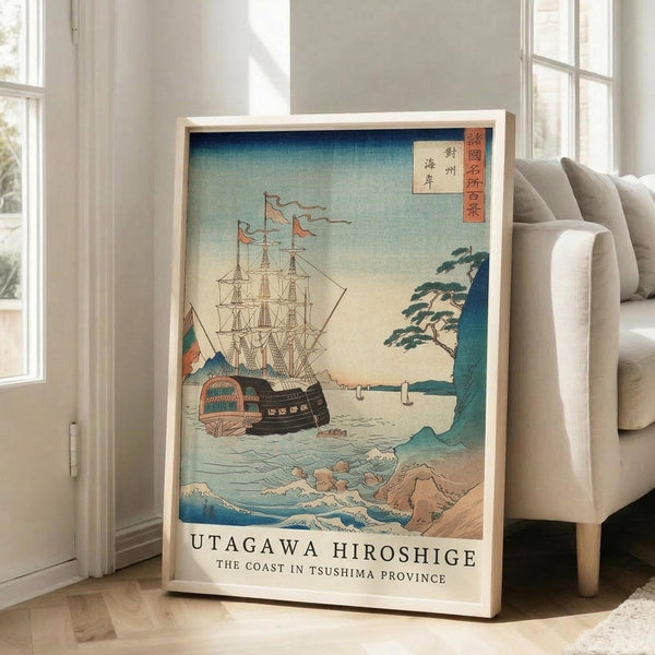 May include: Framed Japanese woodblock print reproduction leaning against white wall next to beige sofa in bright room with windows. Artwork shows sailing ship with tall masts and colorful flags on blue ocean waters near rocky coastline with green pine tree. Traditional ukiyo-e art style with gradient blue sky, white foam waves, and Japanese text characters in red cartouches. Light wood frame, poster appears medium to large size.