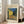 May include: Framed poster of Van Gogh's Cafe Terrace at Night painting showing yellow building with outdoor seating under starry blue sky, displayed on light wooden floor next to beige upholstered sofa in bright room with white walls and windows