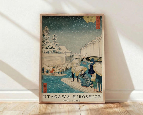 May include: Framed Japanese woodblock print reproduction showing people with umbrellas crossing a snowy ferry scene in winter. The artwork features a turquoise river, falling snow against a dark green sky, traditional buildings, and figures in colorful robes on a wooden boat. Natural wood frame leaning against white wall on light wooden floor with soft window light creating shadows.