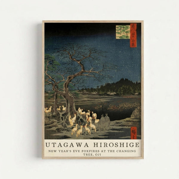 Japanese woodblock print showing foxes with glowing flames gathering beneath a bare tree on a starry night. The scene depicts a moonlit landscape with distant hills and small buildings, rendered in deep blues and earth tones. White and pale yellow foxes create mysterious fires in the foreground, while the skeletal tree branches stretch across a dark sky dotted with stars. The artwork is displayed in a light wooden frame against a white wall, with Japanese text cartouches in the upper right and l
