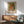 May include: Framed art print featuring a young girl with blonde hair wearing a white dress with red vest, painted in impressionist style with soft brushstrokes and green background. Print displays "PIERRE AUGUSTE exhibition" text and "Claude Renoir" credit. Shown hanging above modern wooden credenza with white ceramic lamp, next to sage green upholstered armchair with wooden frame in bright, naturally lit room.