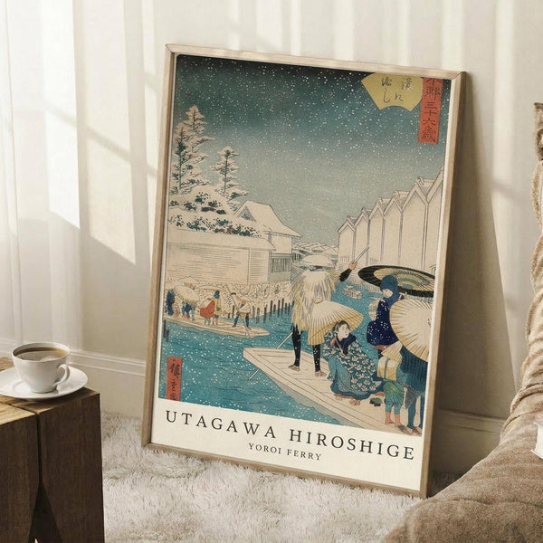 May include: Framed Japanese woodblock print reproduction leaning against white wall corner on fluffy white rug. Shows snowy winter scene with people carrying umbrellas crossing ferry boat, traditional buildings and pine trees in background. Blue-green gradient sky with falling snow, red cartouches with Japanese text in upper corners. White matting, light wood frame. Coffee cup on wooden block nearby, beige throw blanket visible at edge.