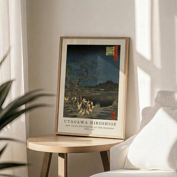 May include: Framed Japanese woodblock print showing foxes gathered under a bare tree at night with starry blue sky, displayed on round wooden side table next to beige upholstered chair. Print features traditional ukiyo-e style with muted earth tones and deep blues. Natural lighting creates soft shadows across white wall background with plant leaves visible in corner.