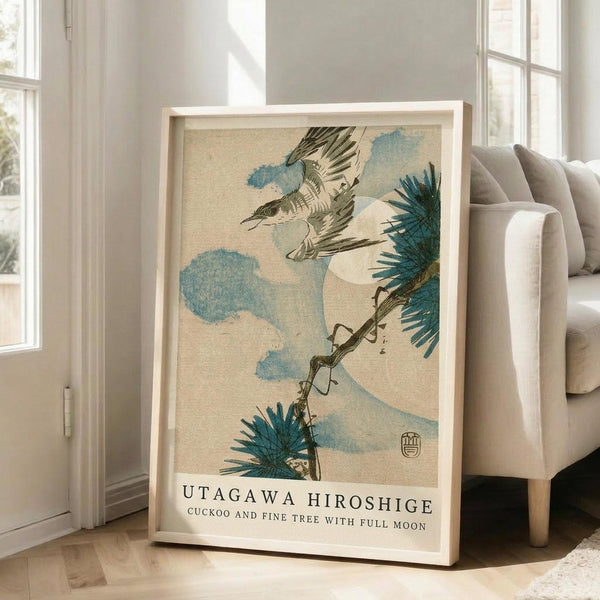 May include: Framed Japanese woodblock print featuring a flying cuckoo bird with outstretched wings above pine tree branches, rendered in muted blues and earth tones on cream background. Light wood frame leans against white wall next to beige upholstered chair in bright, minimalist living room with large windows and natural lighting.