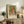May include: Framed exhibition poster featuring Renoir's 1904 painting of a young girl with blonde curls in a white dress with coral accents, set against a soft green background. The cream-colored poster displays "PIERRE AUGUSTE exhibition" text below the artwork. Scene shows the framed print leaning against a white wall on a rustic wooden sideboard, surrounded by neutral decor including a fern plant, ceramic vase, stacked books, and beige upholstered furniture.