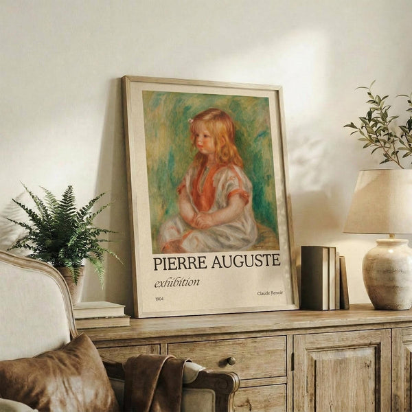 May include: Framed exhibition poster featuring Renoir's 1904 painting of a young girl with blonde curls in a white dress with coral accents, set against a soft green background. The cream-colored poster displays "PIERRE AUGUSTE exhibition" text below the artwork. Scene shows the framed print leaning against a white wall on a rustic wooden sideboard, surrounded by neutral decor including a fern plant, ceramic vase, stacked books, and beige upholstered furniture.