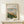 May include: Framed reproduction of Renoir's impressionist painting showing a village view with terracotta buildings nestled among lush green foliage under a cloudy sky. The print has cream matting in a light wood frame, displayed on a pale wooden shelf beside a small wrapped gift with twine and greenery. Soft natural lighting creates gentle shadows on the white wall.