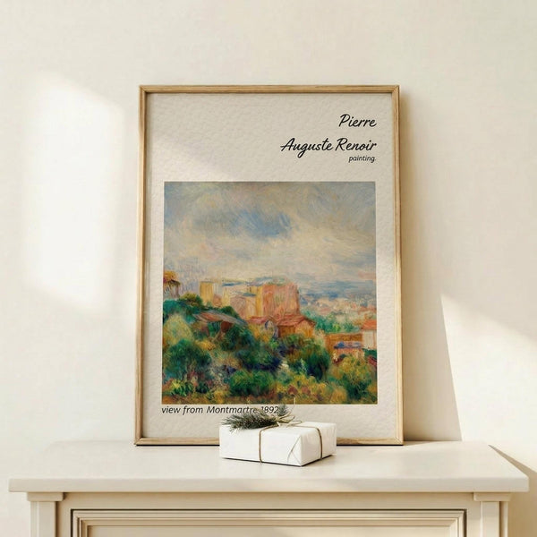 May include: Framed reproduction of Renoir's impressionist painting showing a village view with terracotta buildings nestled among lush green foliage under a cloudy sky. The print has cream matting in a light wood frame, displayed on a pale wooden shelf beside a small wrapped gift with twine and greenery. Soft natural lighting creates gentle shadows on the white wall.