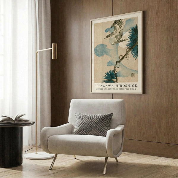 May include: A framed Japanese woodblock print featuring a cuckoo bird in flight among pine branches and blue clouds against a beige background, displayed on wood paneling above a cream upholstered armchair with geometric pillow. Modern floor lamp and side table with open book complete the neutral-toned reading nook scene.