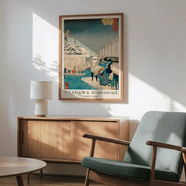 May include: Framed Japanese woodblock print featuring a snowy winter scene with figures crossing water by boat, displayed above a wooden sideboard in a modern living room. The artwork shows traditional architecture, snow-covered trees, and people with umbrellas in teal and earth tones. Scene includes a mint green mid-century armchair, white ceramic lamp, and natural wood furniture with warm sunlight streaming across white walls.