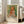 May include: Framed art print leaning against white wall next to beige sofa in bright room with large windows. Print features soft pastel portrait of young girl with blonde hair in orange and white dress against green background. Exhibition-style poster with "PIERRE AUGUSTE" text below painting and "Claude Renoir 1904" attribution. Light wood frame complements neutral living room décor with natural lighting.