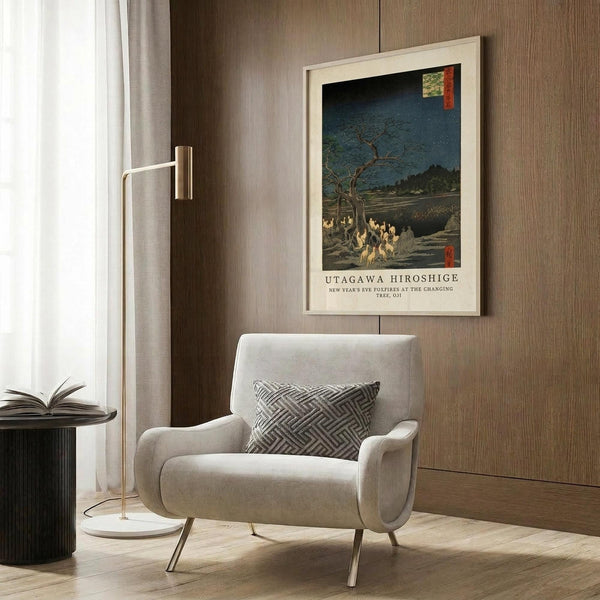 May include: Framed Japanese woodblock print showing foxes gathered under a bare tree at night with blue sky, displayed on wood paneled wall above beige upholstered armchair with geometric patterned pillow, brass floor lamp and dark round side table with open book nearby