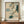 May include: Framed Japanese woodblock print featuring a cuckoo bird in flight with outstretched wings against a soft beige background with blue-green clouds. A pine tree branch with detailed needles extends from the right side, and a pale moon is visible. The artwork displays traditional ukiyo-e style with muted earth tones and teal accents. The frame appears wooden and sits on a mid-century modern credenza alongside a white ceramic vase and stacked books in an elegantly appointed room with whi