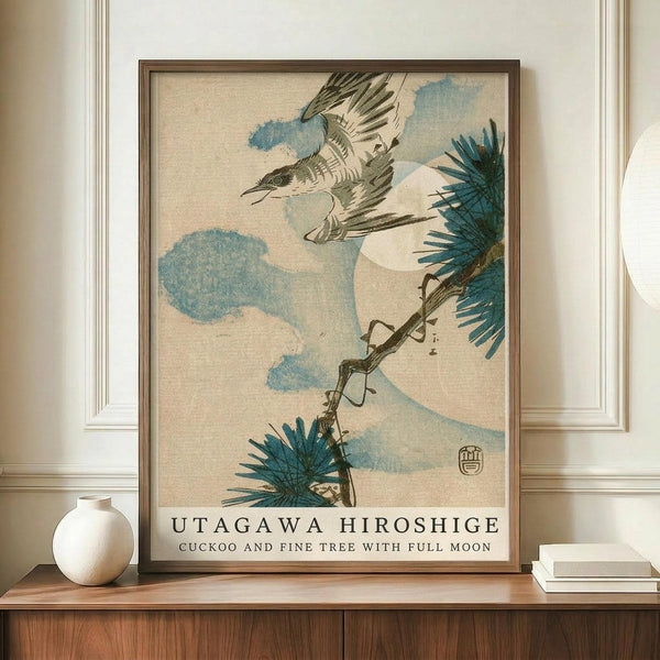 May include: Framed Japanese woodblock print featuring a cuckoo bird in flight with outstretched wings against a soft beige background with blue-green clouds. A pine tree branch with detailed needles extends from the right side, and a pale moon is visible. The artwork displays traditional ukiyo-e style with muted earth tones and teal accents. The frame appears wooden and sits on a mid-century modern credenza alongside a white ceramic vase and stacked books in an elegantly appointed room with whi