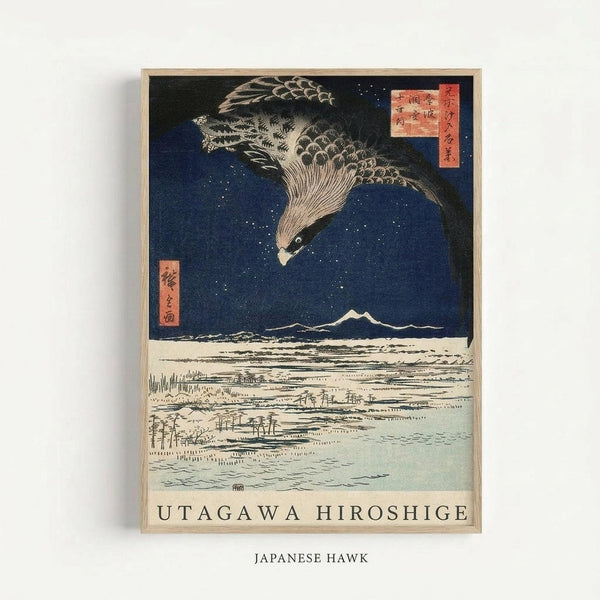 Framed Japanese woodblock print featuring a large hawk with detailed feathers diving through a starry dark blue night sky above a snowy winter landscape. Mount Fuji appears as white peaks in the distance, with a village and bare trees visible in the foreground. Traditional red Japanese text cartouches are positioned in the upper corners. The artwork displays classic ukiyo-e style with bold colors and intricate linework, presented in a light wooden frame against a white wall.
