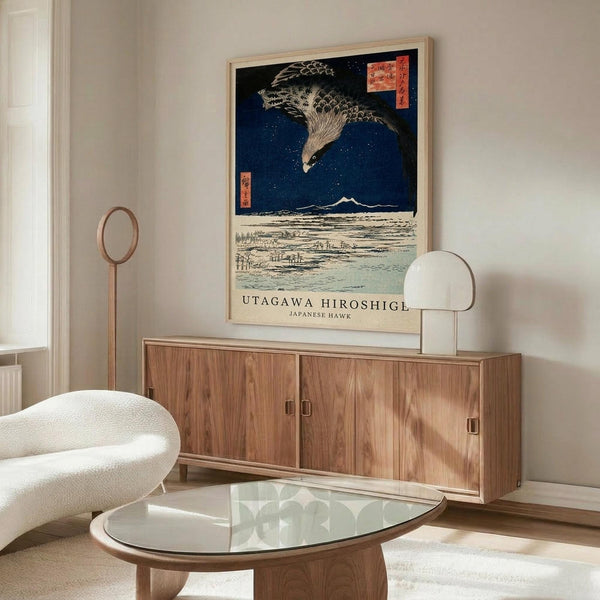 May include: Japanese woodblock print style poster featuring a large hawk with detailed feathers flying over a snowy landscape with mountains and water under a deep blue starry sky, displayed in light wood frame above mid-century modern walnut credenza in minimalist room setting with white curved chair, glass coffee table, and warm natural lighting