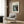 May include: Framed art print of Renoir's impressionist painting showing woman in white dress playing piano, displayed on warm wood paneled wall above beige upholstered armchair with geometric patterned pillow. Modern brass floor lamp and dark round side table with open book complete the cozy reading nook scene with natural lighting from nearby window.