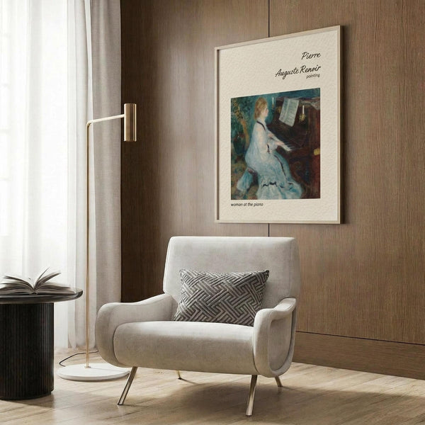 May include: Framed art print of Renoir's impressionist painting showing woman in white dress playing piano, displayed on warm wood paneled wall above beige upholstered armchair with geometric patterned pillow. Modern brass floor lamp and dark round side table with open book complete the cozy reading nook scene with natural lighting from nearby window.