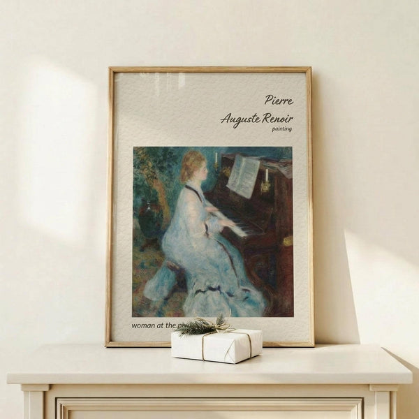 May include: Framed art print of Renoir's impressionist painting showing a woman in a flowing white dress playing piano, displayed on light wood console table with small wrapped gift beside it. The reproduction features soft brushstrokes in blues, whites and earth tones within a thin wooden frame against cream walls.