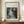 May include: Framed reproduction of Renoir's impressionist painting showing a blonde woman in a flowing white dress playing piano, displayed on a wooden sideboard next to a cream ceramic vase and stacked books against pale walls with white molding in an elegant interior setting.