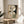 May include: Abstract geometric artwork in wooden frame displayed on round wooden side table next to beige upholstered chair. Art features overlapping circles, grid patterns, and linear elements in neutral tones of cream, black, and sage green. Soft natural lighting creates shadows across white wall. Plant leaves visible in corner of minimalist interior scene.