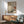 May include: Japanese-inspired artwork displayed in modern living room with mint green mid-century armchair, wooden sideboard, and white table lamp. The framed print features geometric composition with red sun, pine branch, and neutral earth tones in traditional ukiyo-e style. Warm sunlight streams across white walls creating dramatic shadows on the minimalist scandinavian interior.