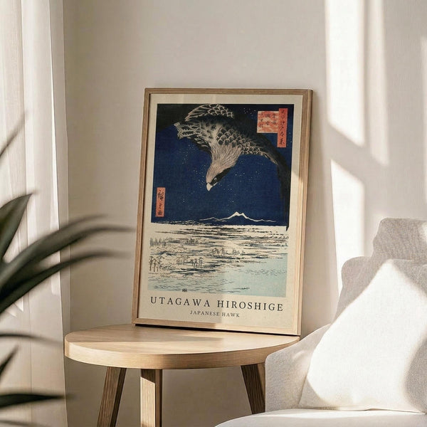 May include: Framed Japanese woodblock print featuring a large hawk with detailed feathers soaring over a snowy landscape with Mount Fuji in the background under a deep blue starry sky. The artwork displays traditional ukiyo-e style with red text cartouches. Shown in a thin wooden frame on a round wooden side table next to a beige upholstered chair, with a plant visible in soft natural lighting.