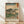 May include: Framed Japanese woodblock print reproduction leaning against white wall on light wood floor. Features Mount Fuji in background with large pine trees, small figures walking along path, and traditional buildings. Soft blue-green sky gradates to warm peachy tones. Red text cartouches in upper corners with Japanese characters. Natural wood frame surrounds artwork. Gentle shadows cast across white wall from natural lighting.