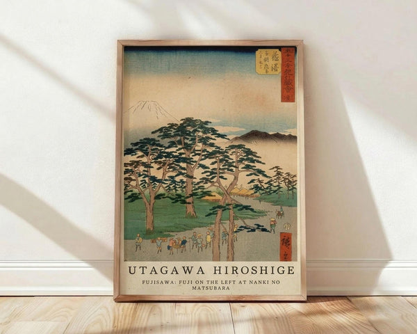 May include: Framed Japanese woodblock print reproduction leaning against white wall on light wood floor. Features Mount Fuji in background with large pine trees, small figures walking along path, and traditional buildings. Soft blue-green sky gradates to warm peachy tones. Red text cartouches in upper corners with Japanese characters. Natural wood frame surrounds artwork. Gentle shadows cast across white wall from natural lighting.
