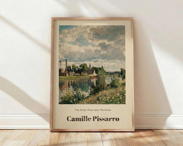 May include: Framed art print of an impressionist landscape painting featuring a river scene with cloudy blue-gray sky, green trees, and buildings reflected in water. The poster has cream-colored matting with "The River Oise near Pontoise" and "Camille Pissarro" text below the artwork. Natural wood frame sits against white wall on light wooden floor with soft window light casting shadows.