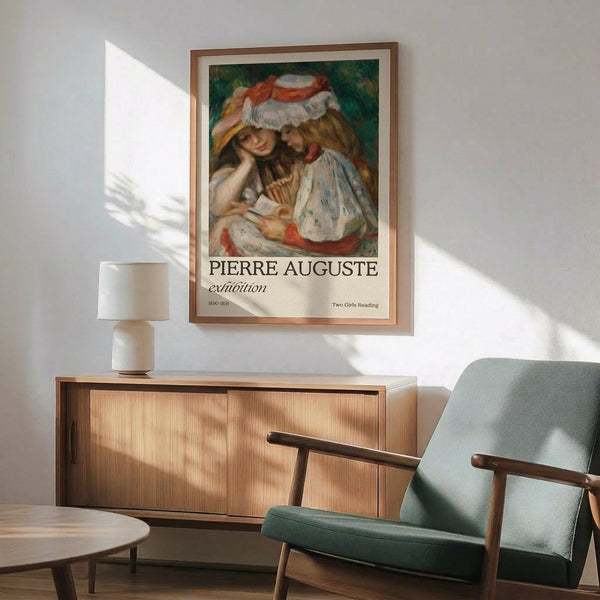 May include: Framed art print hanging above wooden sideboard with white ceramic lamp in modern living room. Print shows two young girls in white dresses and wide-brimmed hats reading together, painted in soft impressionist style with warm colors. Exhibition poster format with "Pierre Auguste" text below artwork. Mint green upholstered armchair with wooden frame visible in foreground. Warm natural lighting creates shadows on white wall.