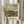 May include: Person in cream sweater holding framed art print showing impressionist landscape painting with river, village buildings, and cloudy sky in muted blues, greens and earth tones. Light wood frame against white curtained window with dried pampas grass stems visible to left side.