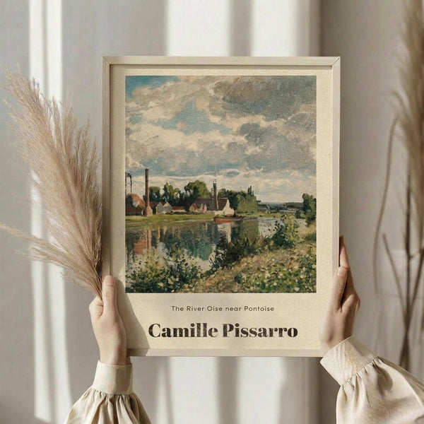 May include: Person in cream sweater holding framed art print showing impressionist landscape painting with river, village buildings, and cloudy sky in muted blues, greens and earth tones. Light wood frame against white curtained window with dried pampas grass stems visible to left side.