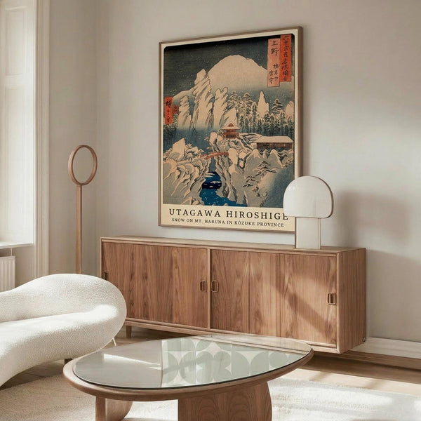 May include: Japanese woodblock print artwork displayed in modern living room setting, featuring snow-covered mountain landscape with traditional buildings and blue water. Framed print hangs above wooden sideboard with glass top coffee table, white upholstered chair, and minimalist floor lamp. Cream walls and natural lighting create serene, contemporary interior design aesthetic.