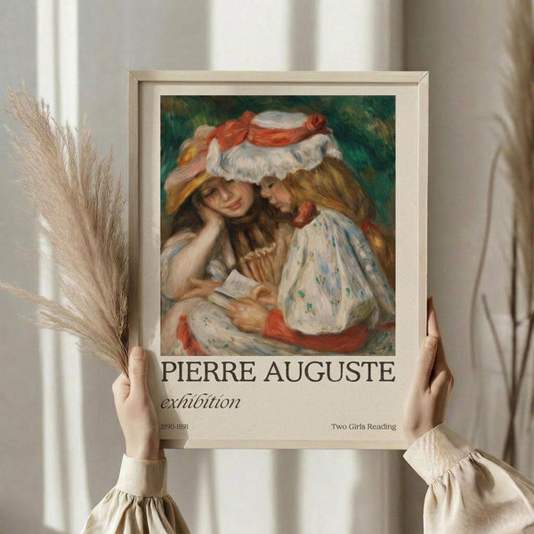 May include: Hands holding a framed art poster featuring two young girls in white dresses and red-ribboned hats reading together, painted in soft impressionist style with warm peachy tones and green background. Exhibition poster format with cream matting in light wood frame. Dried wheat stalks visible beside the frame against white curtained window background.