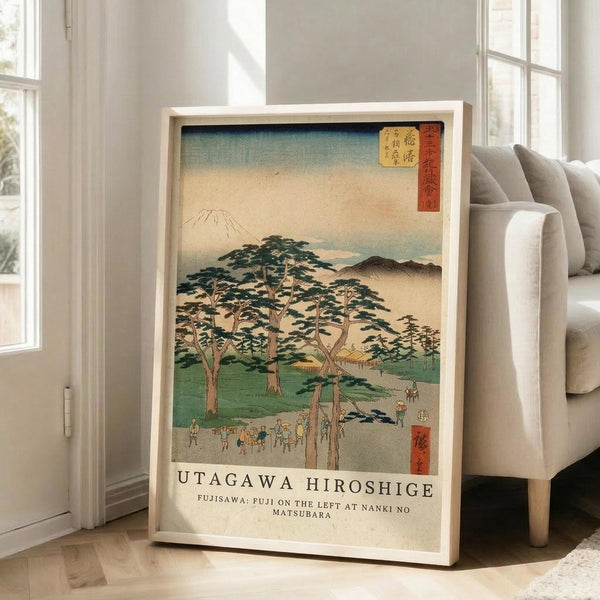 May include: Framed Japanese woodblock print artwork displayed in bright living room with beige sofa and large windows. Print features Mount Fuji in background, tall pine trees, small figures on path, and traditional landscape in muted greens, blues, and earth tones. Light wood frame rests against white wall on hardwood floor.