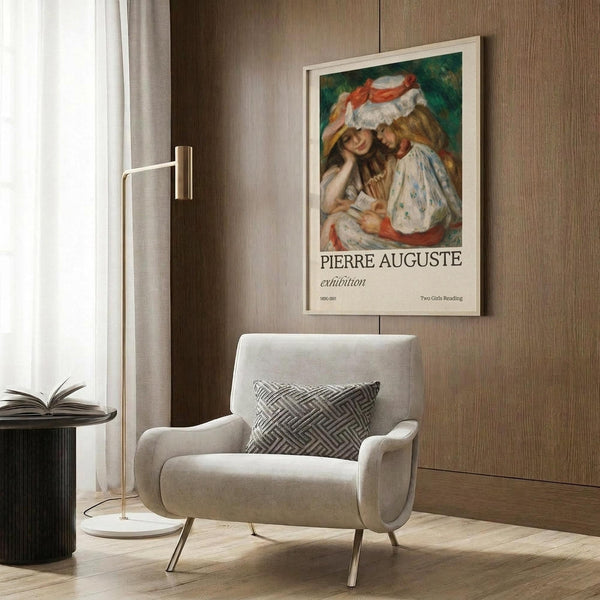 May include: Renoir exhibition poster featuring two young girls reading together, one wearing a white hat with red ribbon. Framed print hangs on wood-paneled wall above cream upholstered armchair with geometric patterned pillow. Modern brass floor lamp and dark round side table with open book visible. Warm neutral interior with curtained window.