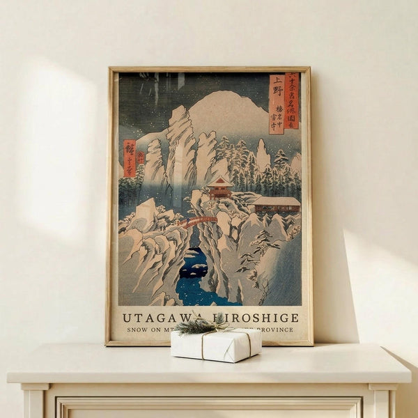 May include: Framed Japanese woodblock print showing snow-covered mountains, traditional buildings, and a blue stream in winter landscape. Print features Japanese text cartouches and muted colors typical of ukiyo-e art style. Light wood frame sits on pale wooden surface next to small white wrapped gift with natural twine and evergreen sprig against cream wall.