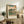 May include: Framed Japanese woodblock print reproduction featuring Mount Fuji in background with traditional pine trees, small figures, and mist-covered landscape in muted greens, blues and earth tones. Light wood frame displayed on rustic wooden sideboard alongside fern plant, ceramic vase, and stacked books in neutral-toned living room setting.